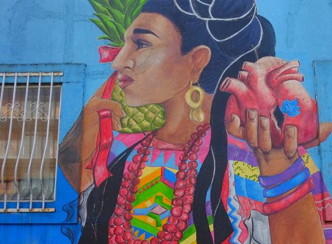 part of a mural, Dance the Dance My Girl by Ari de la Mora and Irving Cato in Vancouver,  A woman in traditional clothing and jewelry, holding a very realistic looking heart and a pineapple.