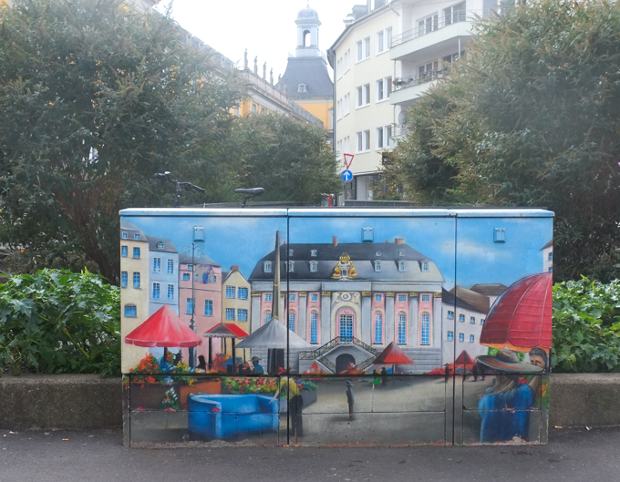 sidewalk utility box in Bonn Germany,painted with a city scene, old city hall, altes rathsaus, a pink and white building with black roof dating from the 18th century