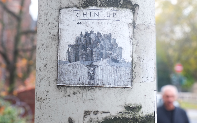 graffiti paste up with text that says chin up, picture of person with backpack on, peering over a fence at a large city