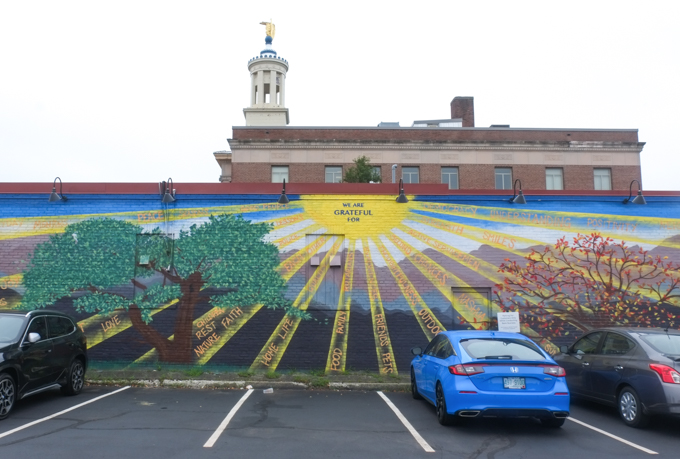 mural from Positive Street Art, What we are grateful for, kindness, family, nature,pets, outdoors, faith, second chances, words are written on rays of the sun that are shining through 4 trees, one for each season,