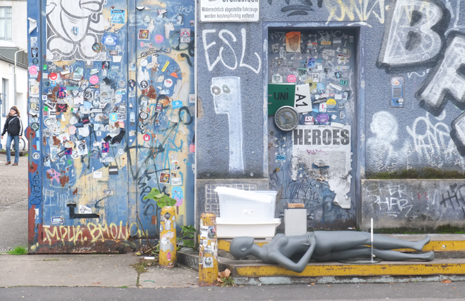 urban art, street art, in Cologne Germany, near Ehrenfeld station, grey mannequin lying on ground outside door that is covered with graffiti, wall beside door is also covered