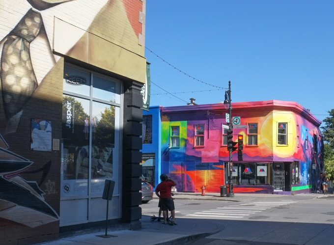 person waiting to cross Blvd Saint Laurent, at Blvd Saint Laurent and Rue Marie-Anne, 2 buildings on opposite corners, each with a street art mural on it