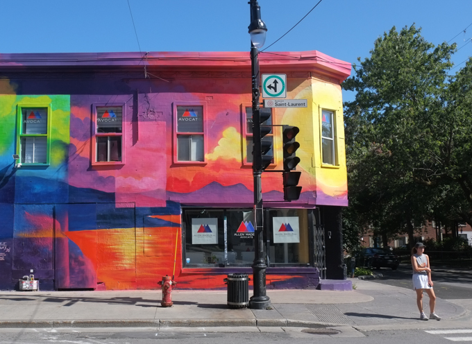 on the side of a building, part of Izabelle Duguay's “Toucher L'horizon” with its pink/blue mountain rising above the orange water.  water is reflecting the setting sun