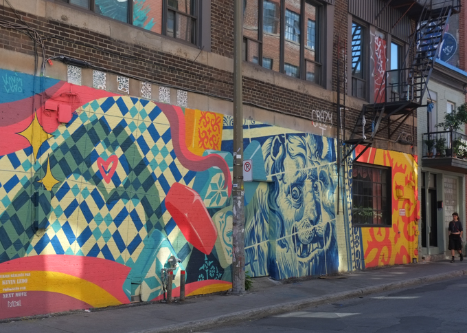mural by Kevin Ledo, blue and white face of a lion flanked by patterns, orange and yellow on one side,  blue and white squares and rectangles on the other class=