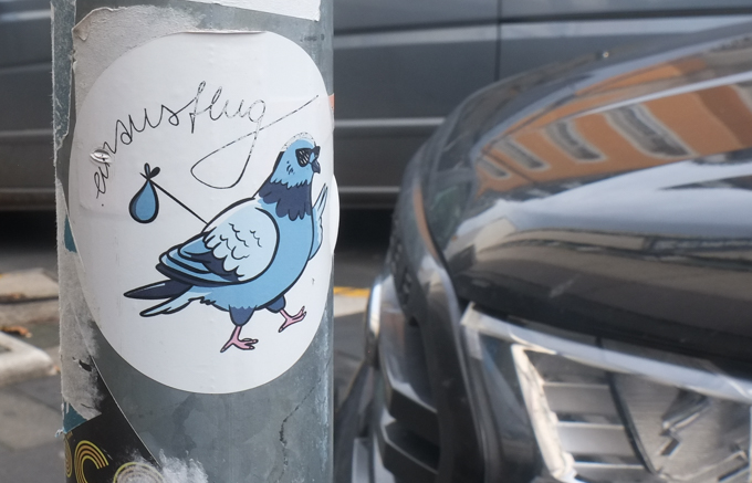graffiti slaps with picture of a pigeon, cursive text says einsusflug, pigeon is wearing sunglasses and carrying asack on a stick over its back