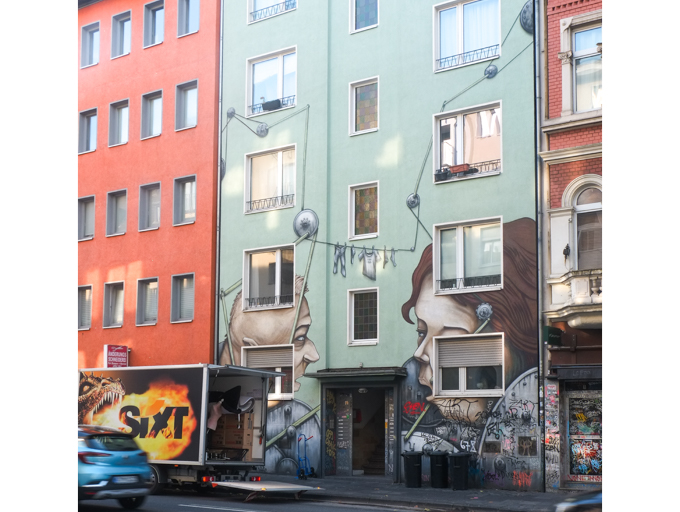mural on front of mint green coloured building, by the Cologne-based artist duo Debug Visuals, consisting of Sebastian Karbowiak and Pierre Galic.  The title is Neighbours, two large faces talking to each other