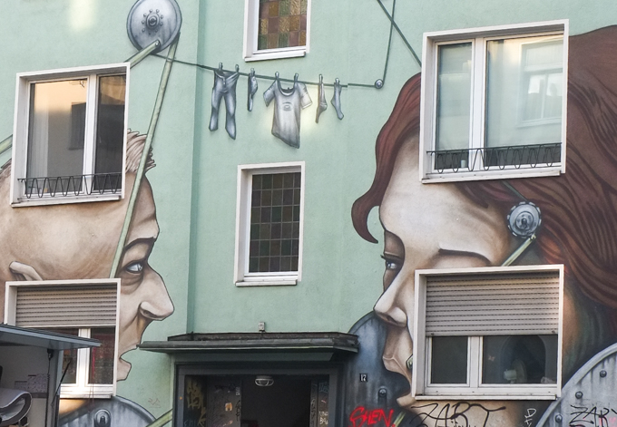 part of mural on front of mint green coloured building, by the Cologne-based artist duo Debug Visuals, consisting of Sebastian Karbowiak and Pierre Galic.  The title is Neighbours, two large faces talking to each other