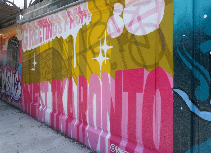 Greetings from West Toronto is the text in this white, gold, and pink mural by Rage Williams. 