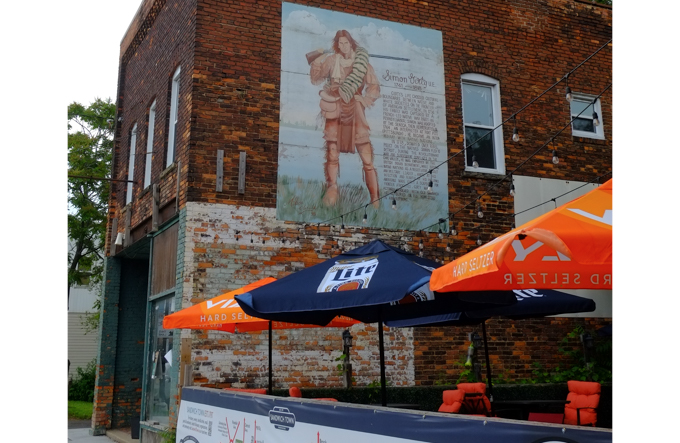 mural about Simon Ginty, picture of him along with many words describing his life on the side of a brick building, above a restaurant patio with blue and orange umbrellas