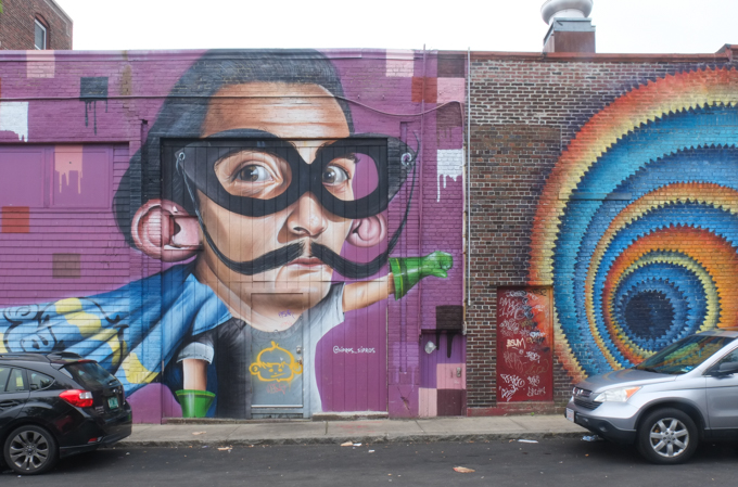 young Dali with long curly mustache, oversized black glasses, and a cape like superman in a mural by Sipros, in Salem Massachusetts
