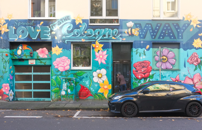 LOVE, It' the Cologne way  in three-dimensional blue letters and cursive script. A bee flies past as a toucan  watches over the garage entrance.  The building has become a garden of flowers including pink hibiscus, pink and red roses, yellow stars, and a white daisy.  mural painted by Henning Hüttner