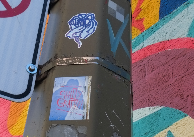 two graffiti stickers, one is a small abstract drawing, the other says shitty graffiti