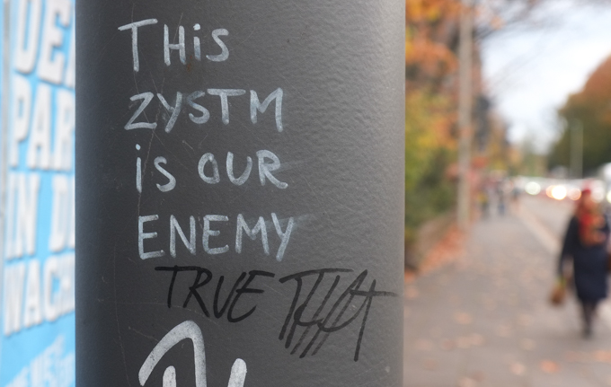 graffiti pasteup with text, the zystem is the enemy, written in white. someone has added, in black, true that