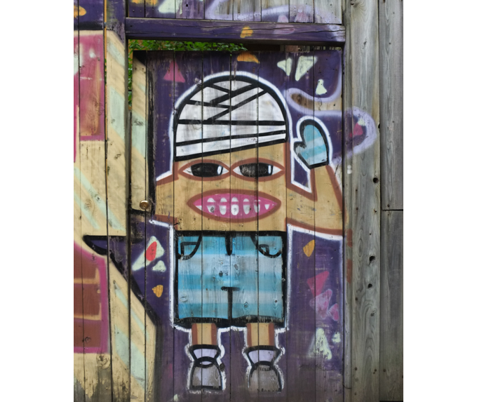 painting on a wood fence, probably male character with head wrapped in white bandages, blue shorts up to chin, big mouth,