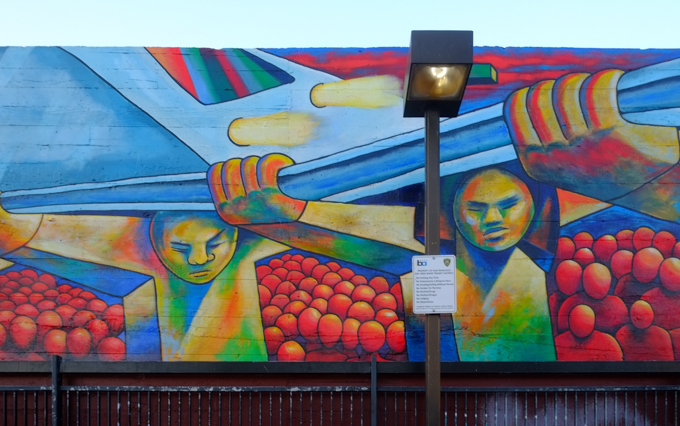 part of a mural at 24th street Bart station, a train is running on elevated tracks that are held up by men, faceless red people wait under the tracks, colourful city buildings in the background