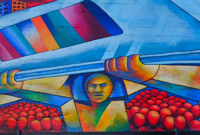 part of a mural at 24th street Bart station, a train is running on elevated tracks that are held up by men, faceless red people wait under the tracks, colourful city buildings in the background