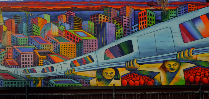 part of a mural at 24th street Bart station, a train is running on elevated tracks that are held up by men, faceless red people wait under the tracks, colourful city buildings in the background, originally painted in 1975 by Michael Rios, Richard Montes and Tony Machado, and updated in 2026