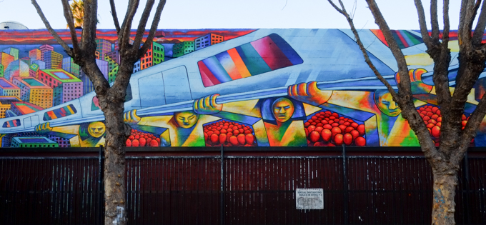 part of a mural at 24th street Bart station, a train is running on elevated tracks that are held up by men, faceless red people wait under the tracks, colourful city buildings in the background