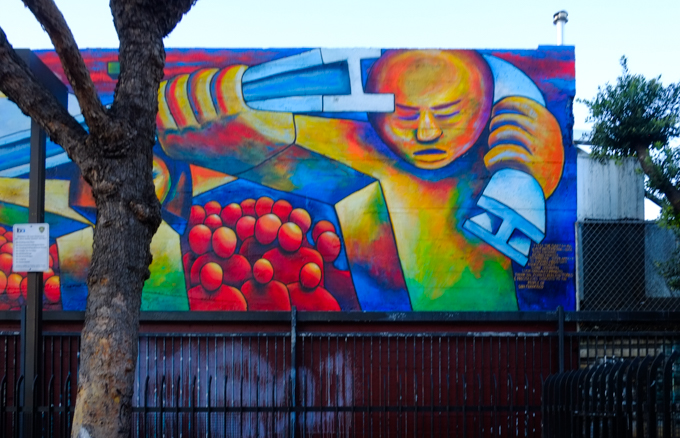 part of a mural at 24th street Bart station, a train is running on elevated tracks that are held up by men, faceless red people wait under the tracks, colourful city buildings in the background, originally painted in 1975 by Michael Rios, Richard Montes and Tony Machado, and updated in 2026