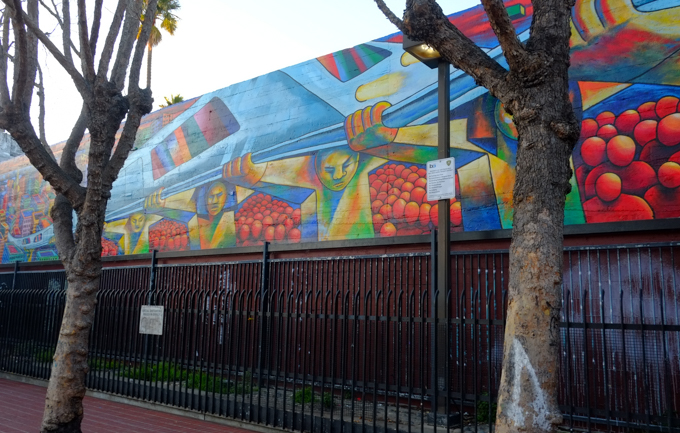 part of a mural at 24th street Bart station, a train is running on elevated tracks that are held up by men, faceless red people wait under the tracks, colourful city buildings in the background, originally painted in 1975 by Michael Rios, Richard Montes and Tony Machado, and updated in 2026