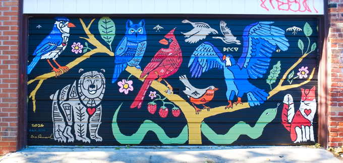 street art mural on garage door in Miriam Garfinkle Lane, a lively collection of birds in a tree - blue jay, robin, owl, cardinal, and more. A smiling grey bear and a fox are under the tree and Canada Geese are flying by. A green snake lurks under the tree.