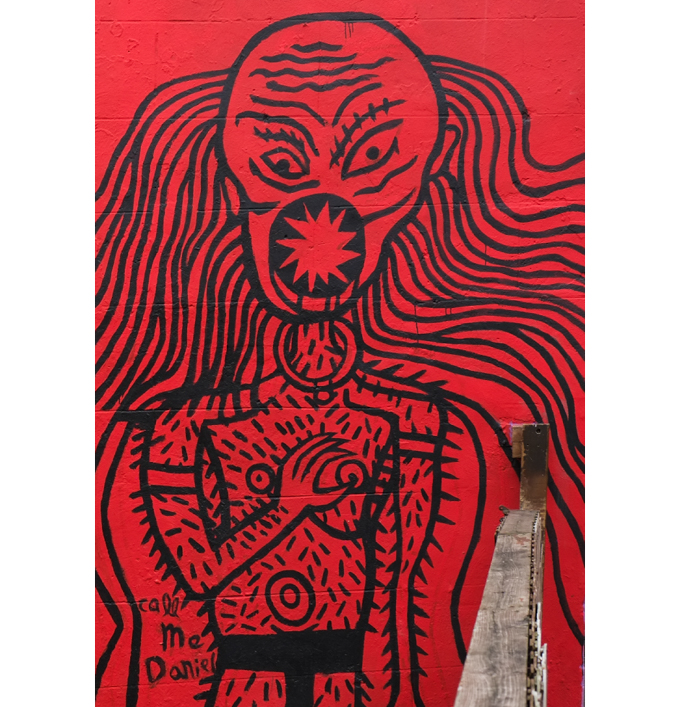 very red background, street art is black line drawing of a person with no hair on top of head but long hair doen the back, open mouth screaming, with text call me Daniel