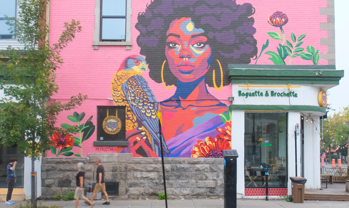 A portrait of a black woman on pink painted brick accompanied by a gorgeous bird and a few flowers in a mural by Melissa Falconer, on side of Baguette & Brochette shop