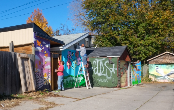 two men working on garages and back fence, surrounded by street art mural on garage door in Miriam Garfinkle Lane,