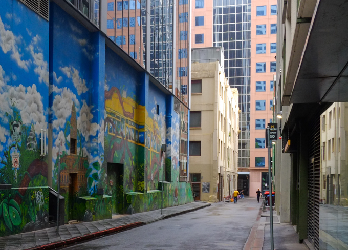 Gallaghers Place in Melbourne, an alley with tall buildings on both sides, murals by Mike Makatron and Conrad Bizjak on one of the walls.