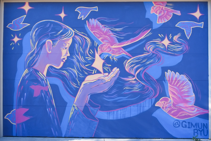 street art mural on garage door in Miriam Garfinkle Lane, by Gimun Ryu, a woman with open hand and pink birds flying towards her