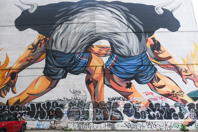 two bodies merged into one at the shoulders, no heads or necks, two silhouettes of bulls behind them, called Liberte, mural painted by Jaz, a k a Franco Fasoli. Arms and legs have tattoos, both wearing blue shorts