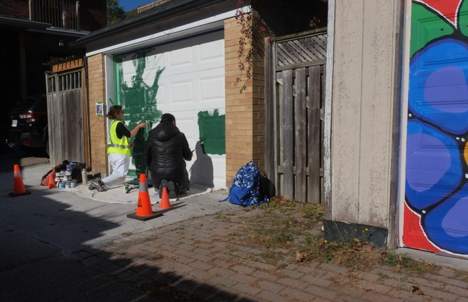 two women paintingstreet art mural on garage door in Miriam Garfinkle Lane,