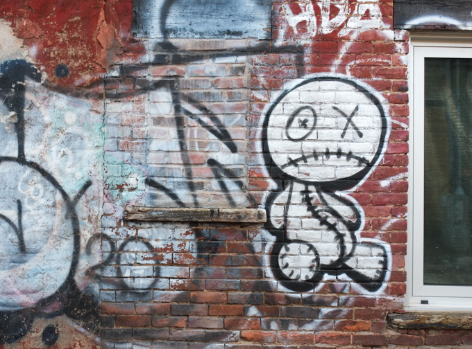 graffiti in Montreal on a red brick wall including a sad character that looks ike a stuffie with stitched mouth and tummy, sad face mouth, one eye is an X and the other eye looks like a button that has been sewn on