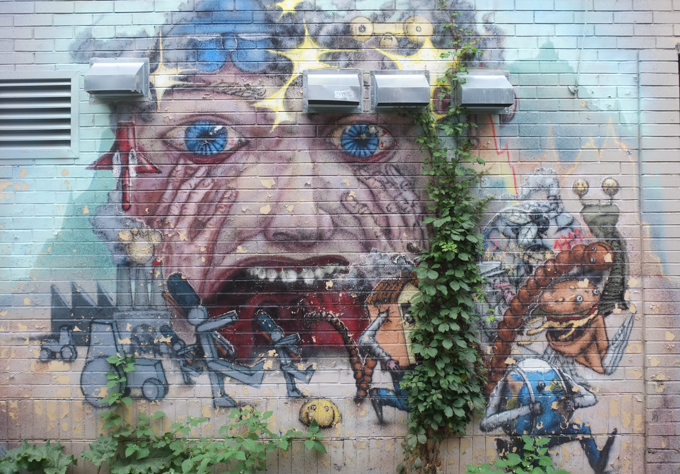 mural by scribe, large face with blood shot blue eyes, wide open red screaming mouth ith teeth, and little characters in front, some faceless ones marching and others running.  runners include a house with legs, a hamburger with legs and an egg shaped globe, also with legs 