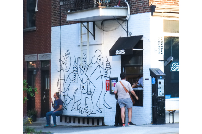 man sitting on bench beside a food shop, with mural on outside wall, line drawing of people with drinks with curly straws, and one dog,